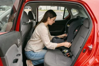 Femme installant un siège auto compact dans une petite voiture citadine rouge, vue de 3/4 depuis l'extérieur du véhicule